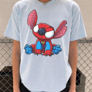 May include: A light blue short-sleeve t-shirt featuring a graphic print of a cartoon character resembling Stitch wearing a red and blue spider-themed costume with a web pattern, large white eyes, and a small yellow spider emblem on the chest.