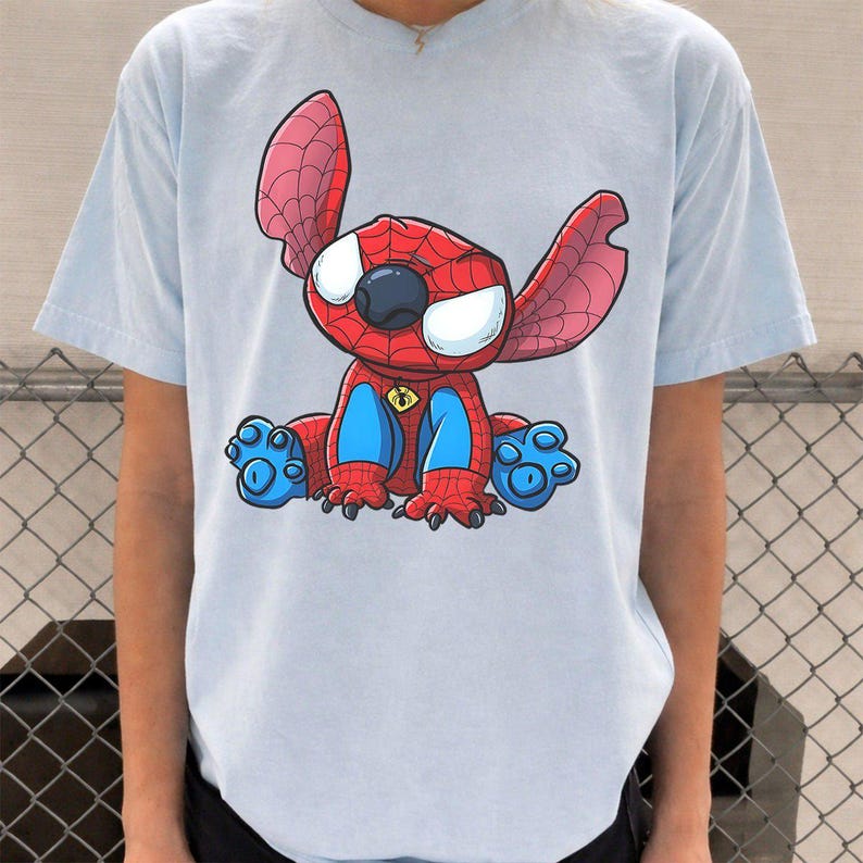 May include: A light blue short-sleeve t-shirt featuring a graphic print of a cartoon character resembling Stitch wearing a red and blue spider-themed costume with a web pattern, large white eyes, and a small yellow spider emblem on the chest.