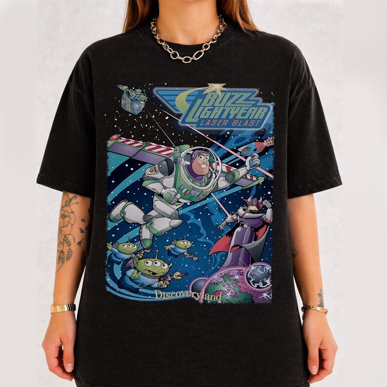 May include: Black t-shirt featuring a colorful graphic of Buzz Lightyear in space, with the text "Buzz Lightyear Laser Blast" and "Discoveryland." The design includes aliens and a villain. The shirt is a relaxed fit.