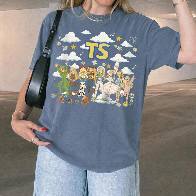 May include: A slate blue short-sleeve t-shirt featuring a graphic print of various animated characters in different outfits, white clouds, yellow stars, and the text TS in large yellow letters. The shirt has a crew neckline and a relaxed fit.