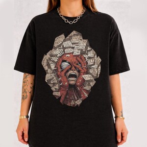 May include: A black short-sleeve t-shirt featuring a graphic print of a red masked character with hands on their head, surrounded by a collage of scattered paper notes with handwritten numbers and mathematical equations in a jagged shape.