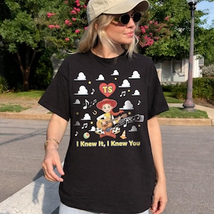 May include: A black short-sleeve t-shirt featuring a graphic of a cowgirl playing a guitar, surrounded by white clouds, musical notes, stars, a heart with the letters TS, and the text I Knew It, I Knew You printed in yellow at the bottom.