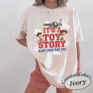 K&ouml;nnte beinhalten: Ein elfenbeinfarbenes Kurzarm-T-Shirt mit einem Grafikdruck von Woody, Buzz Lightyear und Jessie aus Toy Story. Der Text lautet IT'S A TOY STORY BABY JUST SAY YES in roter und blauer Schrift, umgeben von wei&szlig;en Wolken und gelben Sternen.