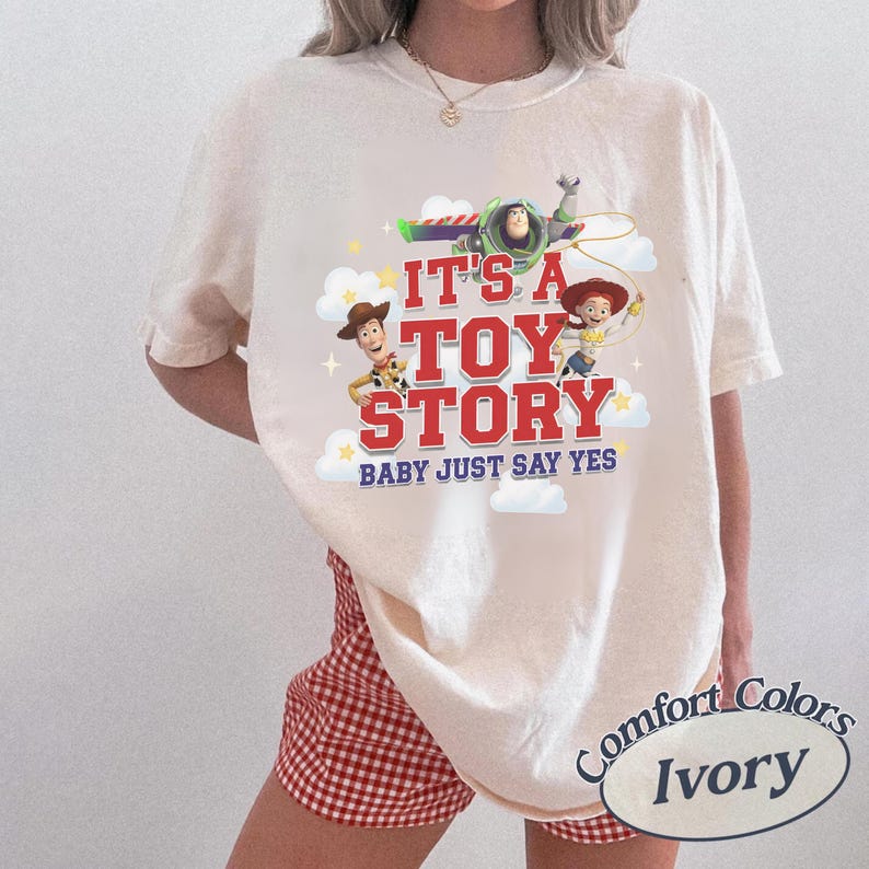 K&ouml;nnte beinhalten: Ein elfenbeinfarbenes Kurzarm-T-Shirt mit einem Grafikdruck von Woody, Buzz Lightyear und Jessie aus Toy Story. Der Text lautet IT'S A TOY STORY BABY JUST SAY YES in roter und blauer Schrift, umgeben von wei&szlig;en Wolken und gelben Sternen.