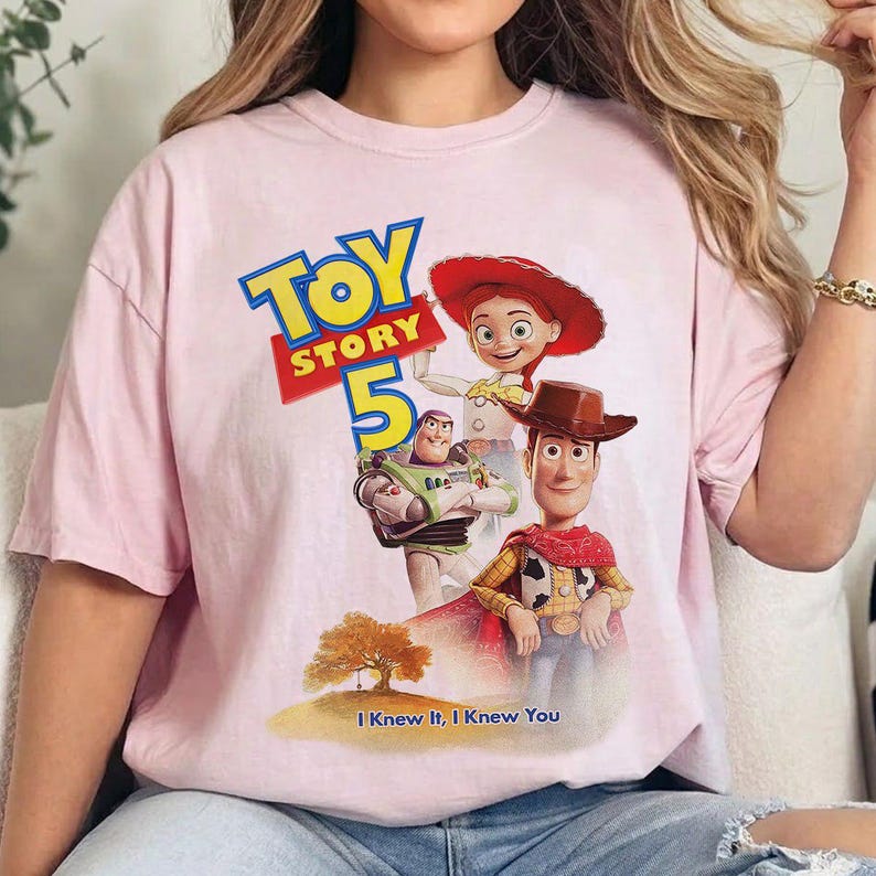 May include: A light pink short-sleeve t-shirt featuring a graphic print of Toy Story characters Woody, Buzz Lightyear, and Jessie. The design includes the text Toy Story 5 and the phrase I Knew It, I Knew You above a small tree illustration.