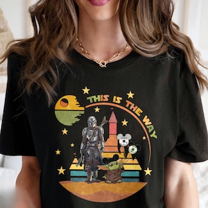 Puede incluir: Camiseta negra con un dise&ntilde;o gr&aacute;fico que presenta el texto "THIS IS THE WAY". El dise&ntilde;o incluye una figura mandaloriana, Baby Yoda y otros elementos tem&aacute;ticos de Star Wars. El dise&ntilde;o est&aacute; rodeado de estrellas.