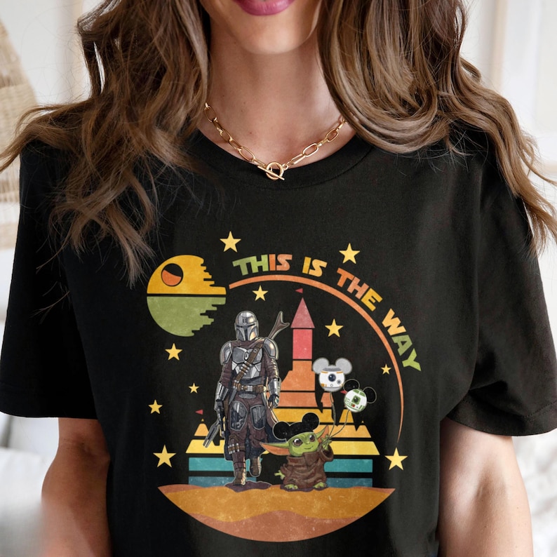 Puede incluir: Camiseta negra con un dise&ntilde;o gr&aacute;fico que presenta el texto "THIS IS THE WAY". El dise&ntilde;o incluye una figura mandaloriana, Baby Yoda y otros elementos tem&aacute;ticos de Star Wars. El dise&ntilde;o est&aacute; rodeado de estrellas.