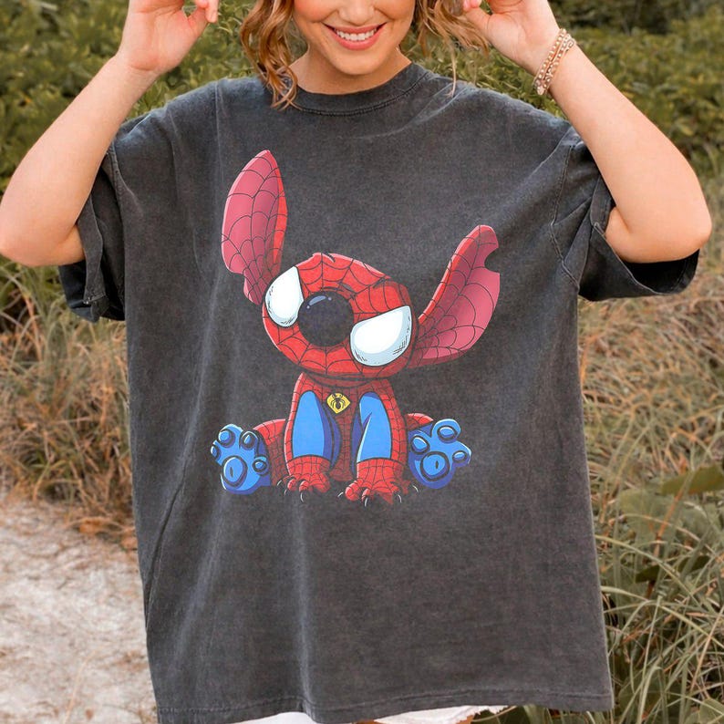 May include: A charcoal gray short-sleeve t-shirt featuring a graphic print of a character with large ears and eyes, wearing a red and blue spider-web patterned suit. The character is sitting down and has blue paws with visible pads.