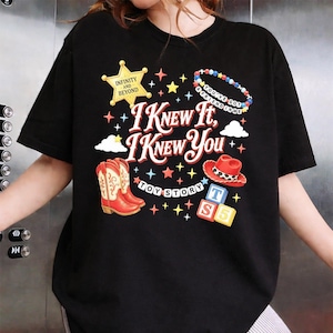 May include: A black t-shirt featuring a graphic print with the text I KNEW IT, I KNEW YOU, TOY STORY, INFINITY AND BEYOND, and YOU'VE GOT A FRIEND IN ME. The design includes a yellow sheriff star, red cowboy boots, a red hat, and colourful blocks.