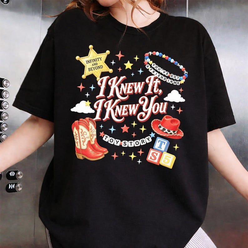 May include: A black t-shirt featuring a graphic print with the text I KNEW IT, I KNEW YOU, TOY STORY, INFINITY AND BEYOND, and YOU'VE GOT A FRIEND IN ME. The design includes a yellow sheriff star, red cowboy boots, a red hat, and colourful blocks.