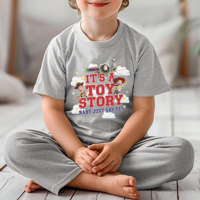 K&ouml;nnte beinhalten: Ein graumeliertes Kurzarm-T-Shirt mit einem Grafikdruck von Buzz Lightyear, Woody und Jessie aus Toy Story. Der Text lautet IT'S A TOY STORY BABY JUST SAY YES, umgeben von wei&szlig;en Wolken und kleinen gelben Sternen auf der Vorderseite des Brustbereichs.