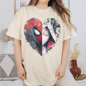 May include: A cream-colored short-sleeve t-shirt featuring a large heart-shaped graphic on the chest. The graphic is split vertically, showing a red Spider-Man mask on the left and a white Spider-Gwen mask on the right, with comic book style details.
