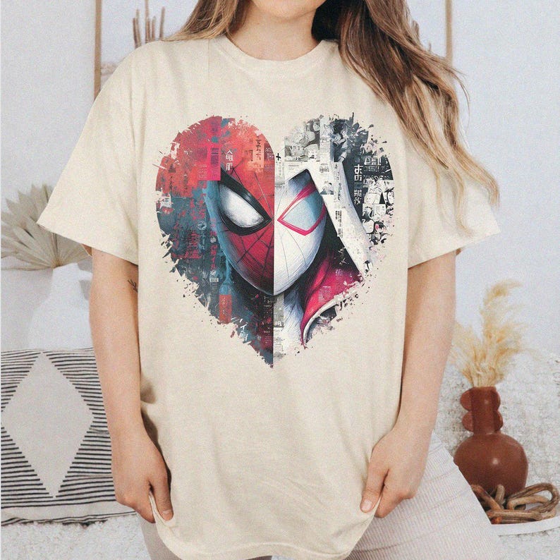 May include: A cream-colored short-sleeve t-shirt featuring a large heart-shaped graphic on the chest. The graphic is split vertically, showing a red Spider-Man mask on the left and a white Spider-Gwen mask on the right, with comic book style details.