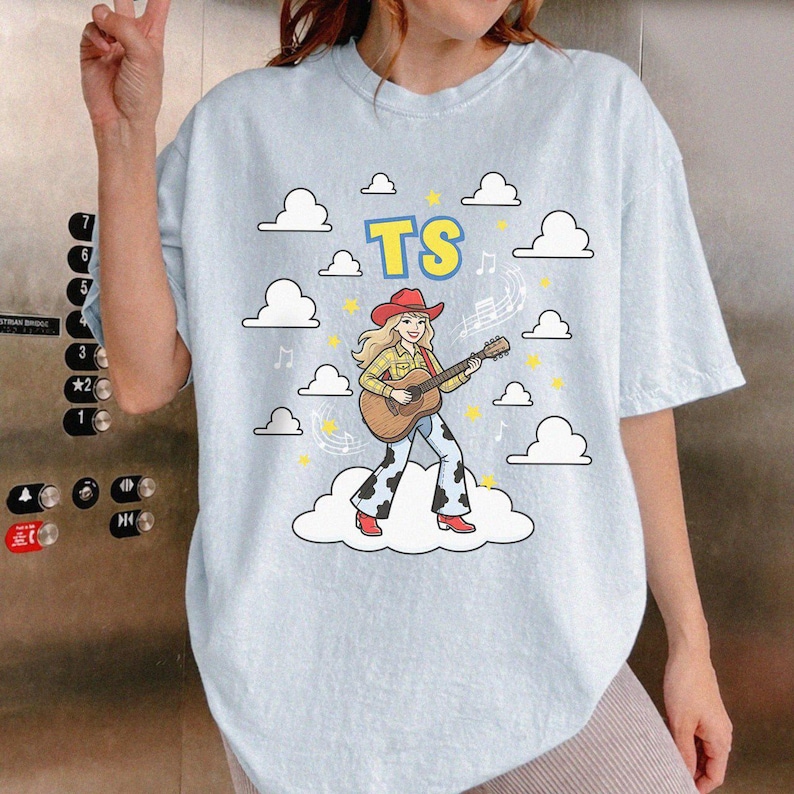 May include: A light blue t-shirt featuring a graphic of a blonde person in a red cowboy hat, yellow plaid shirt, and cow-print trousers playing an acoustic guitar. The design includes white clouds, yellow stars, musical notes, and the letters TS.