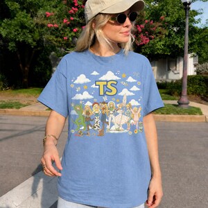 May include: A light blue short-sleeve t-shirt featuring a graphic print of various cartoon characters in different outfits. The text TS is printed in yellow at the top center, surrounded by white clouds, stars, and musical notes on a blue background.