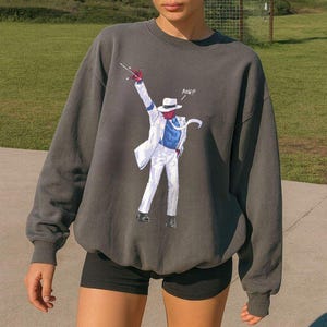 May include: A charcoal gray crewneck sweatshirt featuring a graphic print of a figure in a white suit, blue shirt, and white fedora, striking a dance pose with one arm raised. The graphic includes the text "AOW!!" in white lettering.