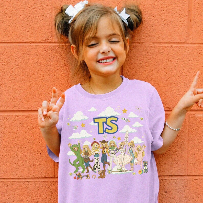 May include: A light purple t-shirt featuring a graphic print of various cartoon characters in a group, with the letters TS in yellow and blue at the top. The design includes white clouds, yellow stars, and small musical notes scattered across the front.