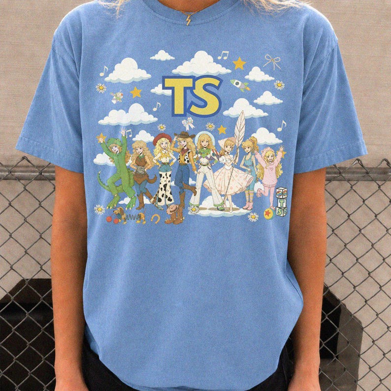 May include: A light blue short-sleeve t-shirt featuring a graphic print with the text TS in yellow. The design includes a group of illustrated characters in various outfits, surrounded by white clouds, yellow stars, musical notes, and small flowers.
