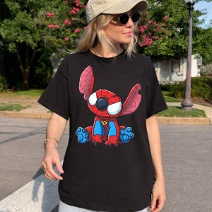 May include: A black short-sleeved t-shirt featuring a graphic print of a cartoon character resembling a blue alien creature wearing a red and blue spider-themed suit with a web pattern, large white eyes, and a small yellow spider emblem on the chest.