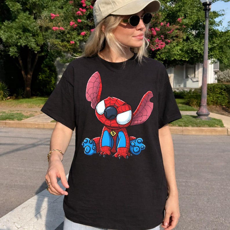 May include: A black short-sleeved t-shirt featuring a graphic print of a cartoon character resembling a blue alien creature wearing a red and blue spider-themed suit with a web pattern, large white eyes, and a small yellow spider emblem on the chest.