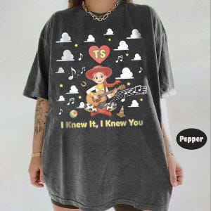 May include: A charcoal gray t-shirt featuring a graphic of a cartoon cowgirl playing an acoustic guitar. The design includes a red heart with the letters TS, white clouds, musical notes, stars, and the text I Knew It, I Knew You in yellow.