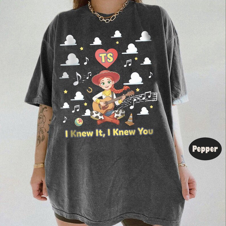 May include: A charcoal gray t-shirt featuring a graphic of a cartoon cowgirl playing an acoustic guitar. The design includes a red heart with the letters TS, white clouds, musical notes, stars, and the text I Knew It, I Knew You in yellow.