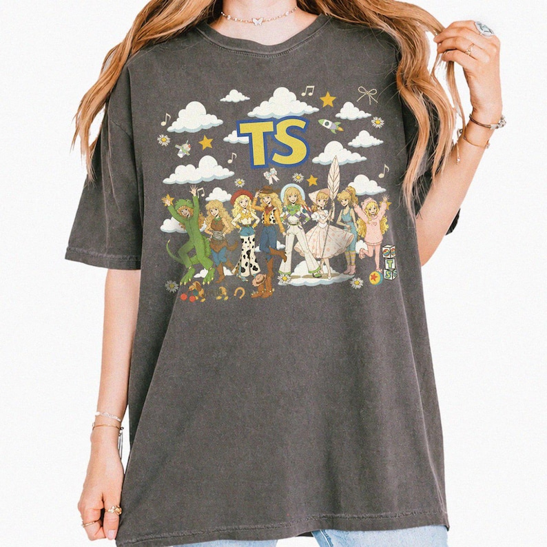 May include: A charcoal gray short-sleeve t-shirt featuring a graphic print of multiple animated characters in various outfits, white clouds, yellow stars, and the letters TS in bold yellow font. The design includes musical notes and small icons.