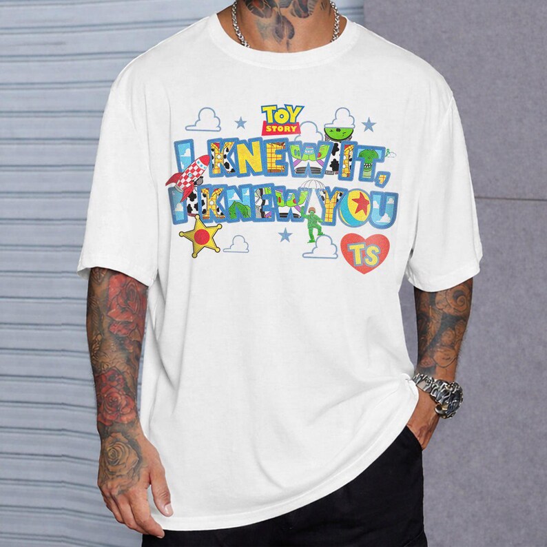 Pode incluir: Uma t-shirt branca de manga curta com uma impress&atilde;o gr&aacute;fica colorida do Toy Story. O design inclui o texto "I KNEW IT, I KNEW YOU" em letras azuis, um cora&ccedil;&atilde;o vermelho com "TS" dentro, uma estrela amarela e v&aacute;rios &iacute;cones.