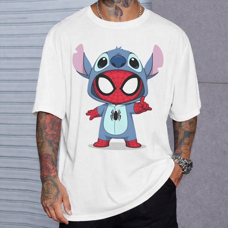 May include: A white short-sleeved t-shirt featuring a graphic print of a blue character wearing a red and blue spider-themed costume with a hood. The character has large eyes, pointed ears, and a small black spider emblem on the chest area.