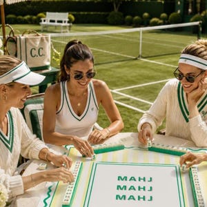 May include: Three people are playing mahjong outdoors. They are wearing white and green tennis-themed outfits. The table has a mahjong board with the word "MAHJ" printed on it. A canvas tote bag with "C.C." printed on it is visible.
