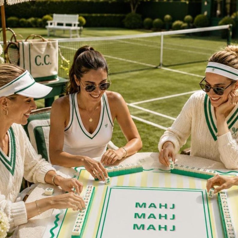 May include: Three people are playing mahjong outdoors. They are wearing white and green tennis-themed outfits. The table has a mahjong board with the word "MAHJ" printed on it. A canvas tote bag with "C.C." printed on it is visible.