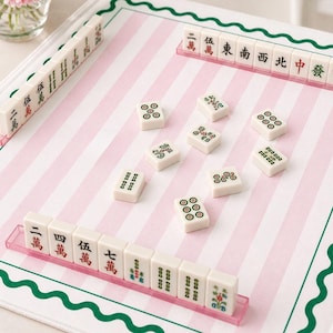 May include: A Mahjong set with ivory-colored tiles featuring red, green, and black symbols and characters. The tiles are arranged on a pink and white striped playing surface with a green border. The set includes racks for holding the tiles.