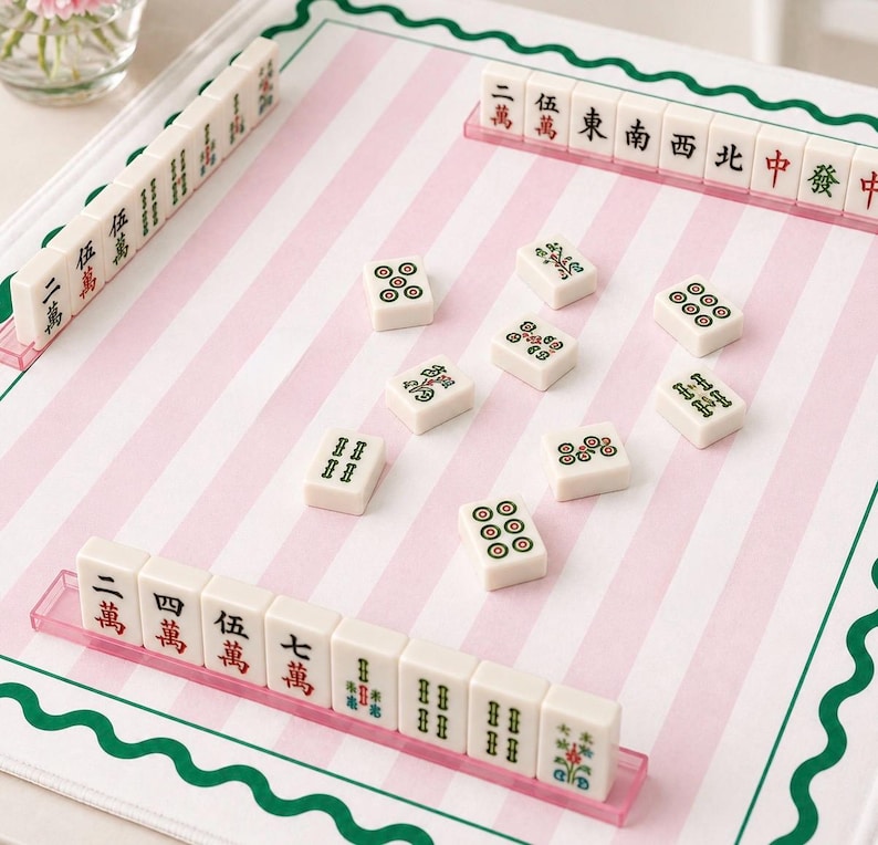 May include: A Mahjong set with ivory-colored tiles featuring red, green, and black symbols and characters. The tiles are arranged on a pink and white striped playing surface with a green border. The set includes racks for holding the tiles.