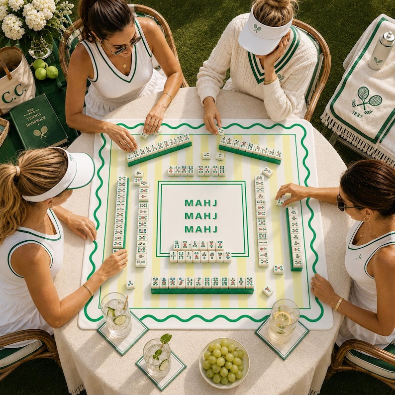 May include: Four people playing Mahjong on a round table. The Mahjong set has green and white tiles and a white board with green and yellow stripes. The table is set with drinks, coasters, and a bowl of grapes. The players are wearing white tennis attire.