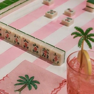 May include: A still life featuring a Mahjong game in progress, with ivory tiles and green accents on a pink and white striped surface. A pink cocktail with a lemon slice and palm tree decoration sits nearby, along with a pink napkin embroidered with a palm tree.