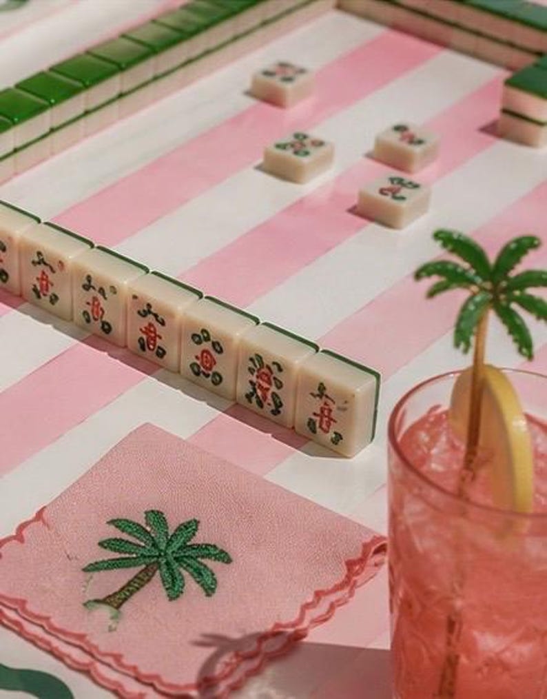 May include: A still life featuring a Mahjong game in progress, with ivory tiles and green accents on a pink and white striped surface. A pink cocktail with a lemon slice and palm tree decoration sits nearby, along with a pink napkin embroidered with a palm tree.
