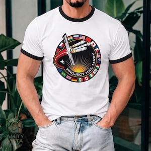 May include: White t-shirt with black trim featuring a circular graphic with the text "PROJECT HAIL MARY". The graphic includes a rocket design, a star, and flags of various countries. The shirt is a crew neck with short sleeves.