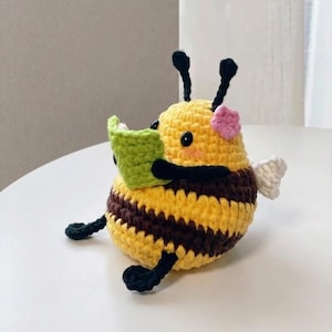 Puede incluir: Figura de abeja de ganchillo, con rayas amarillas y marrones, sosteniendo un libro verde. La abeja tiene antenas, ojos y patas negras, con una flor rosa y alas blancas. Un artículo artesanal.