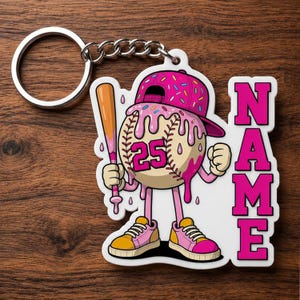 Könnte beinhalten: Ein Baseball-Schlüsselanhänger mit einem Cartoon-Baseball-Charakter, der eine rosa, mit Donuts verzierte Baseballkappe trägt. Die Figur hält einen Schläger und hat die Nummer 25 auf dem Baseball. Das Wort "NAME" wird in rosa Buchstaben angezeigt.