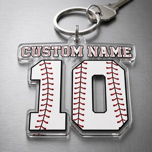 Könnte beinhalten: Transparenter Acryl-Schlüsselanhänger mit dem Text "CUSTOM NAME" über der Zahl 10, gestaltet wie ein Baseball. Die Zahl 10 ist weiß mit roten Nähten, und der Schlüsselanhänger ist an einem silbernen Schlüsselring und einem Schlüssel befestigt.