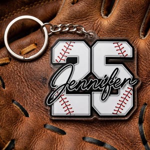 May include: A baseball-themed keychain with the number 25 and the name "Jennifer" in a script font. The keychain features a white background with red stitching details, resembling a baseball. The background is a brown leather baseball glove.