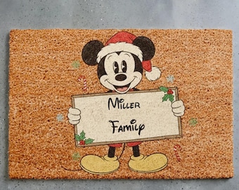 Puede incluir: Un felpudo rectangular de fibra de coco color canela con un gráfico de Mickey Mouse con un gorro de Papá Noel rojo. Mickey sostiene un cartel rectangular con el texto Miller Family en negro, decorado con acebo verde y bayas rojas.