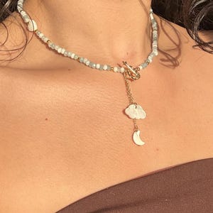 May include: A delicate necklace featuring a beaded strand with white, light blue, and gold accents. A gold clasp secures the necklace, which has a dangling charm of a white cloud and a crescent moon.