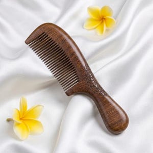 May include: A wooden comb with a curved handle and fine teeth. The comb is a rich brown color with a decorative carved design on the handle. Two yellow flowers are placed near the comb on a white satin surface.