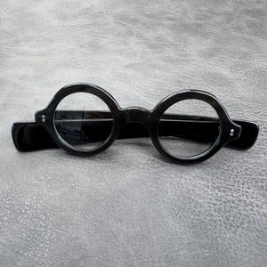 May include: A pair of black eyeglasses featuring circular frames and clear lenses. The frames have a glossy finish with two small silver-toned rivets on each side where the temples connect to the front, showcasing a classic round design.