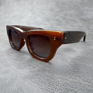 Puede incluir: Gafas de sol con una montura gruesa de color ámbar translúcido y lentes oscuras. La montura presenta una forma de ojo de gato con dos pequeños remaches metálicos de color plateado en cada lado del frente y en las patillas.