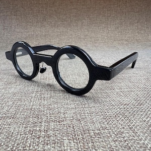May include: Black round-frame spectacles. The frames are thick and have a textured appearance. The lenses are clear. The arms of the spectacles are straight and extend back from the circular frames. The spectacles are set against a neutral background.