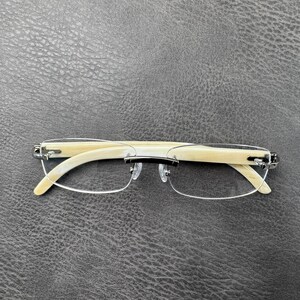 May include: Rimless eyeglasses featuring cream-colored, smooth-textured temples and a silver-tone metal bridge. The lenses are held in place by a thin silver wire frame, and the nose pads are clear, transparent plastic attached to the bridge.