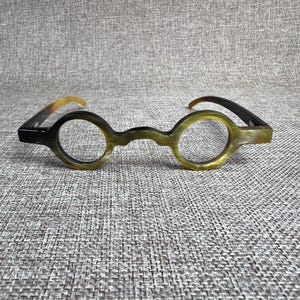 Può includere: Occhiali rotondi dal design unico. Le montature sono un mix di nero e verde oliva, con una texture simile al corno. Le aste sono nere e passano a un colore marrone chiaro. Gli occhiali sono su uno sfondo grigio strutturato.