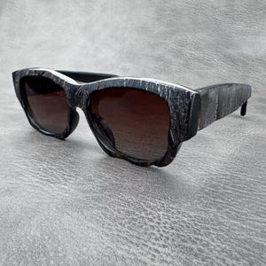 May include: Sunglasses featuring a thick, chunky frame with a dark, textured, marbled pattern in shades of black and grey. The lenses are dark brown and semi-transparent, set within a wide, angular frame design with prominent, sturdy temples.